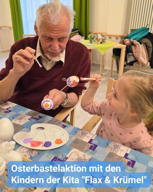 Nahaufnahme eines älteren Mannes mit kurzen, grauen Haaren, der einen burgunderfarbenen Pullover trägt und an einer Bastelaktivität zu Ostern mit einem kleinen Mädchen teilnimmt.  Der Mann sitzt an einem Tisch, hält sorgfältig ein kleines bemaltes Ei sowie einen Pinsel in der Hand. Das Mädchen mit hellbraunen Haaren sitzt ebenfalls am Tisch, hält einen Pinsel und betrachtet aufmerksam ihr Werk. Der Tisch ist mit einer hellblauen Tischdecke in einem gemusterten Design bedeckt, und eine kleine Palette mit verschiedenen Farbnuancen ist sichtbar.  Eine Beschriftung am unteren Rand des Bildes, weist darauf hin, dass es sich um eine Osterbastelstunde mit den Kindern einer Kindertagesstätte namens "Flax & Krümel" handelt. Auf dem Tisch sind zudem weitere Bastelmaterialien sowie einige osterliche Utensilien zu sehen, darunter ein Eierbecher.