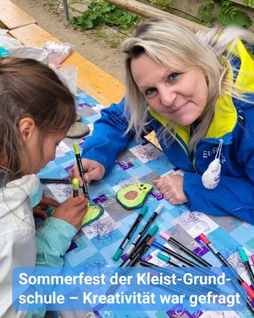 Ein mittlerer Blick fängt ein junges Mädchen und eine erwachsene Frau ein, die an einem Tisch sitzen, der mit einem blau-weißen karierten Tischlaken bedeckt ist. Beide Figuren sind beim Zeichnen auf bemalten Steinen vertieft, die wie Avocados gestaltet wurden. Die Frau, die älter wirkt, trägt eine blaue Jacke mit gelben Akzenten und blickt mit einem dezenten Lächeln in Richtung Kamera.  Das Mädchen befindet sich auf der linken Seite des Bildes, zeigt größtenteils ihren Rücken und die Seite ihres Gesichts. Sie hält ebenfalls einen Marker und zeichnet sorgfältig auf den Stein-Avocado. Eine Sammlung buntfarbiger Marker ist über den Tisch verstreut.  Am unteren Rand des Bildes befindet sich Text, der anzeigt, dass es sich um ein Sommerfest an der Kleist-Grundschule handelt, bei dem Kreativität gefördert wird. Der Hintergrund deutet auf eine Outdoor-Umgebung hin..